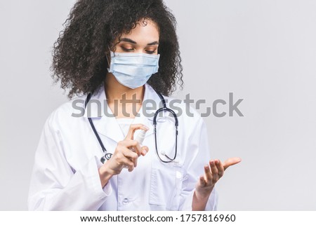 Young african american woman doctor in medical uniform and protective mask using hand sanitizer gel, isolated on grey background. Hand hygiene and antibacterial protection. 