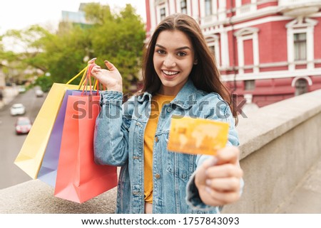 Image of young happy woman in denim wear smiling while holding credit card and shopping bags on city street