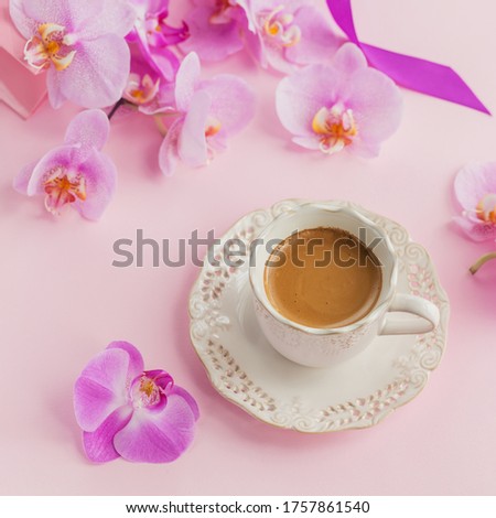 Delicate flatlay composition with morning cup of coffee with milk or cappuccino, pink gift bag and orchid flowers on light pink background. Beautiful breakfast concept