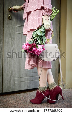 A woman in a romantic dress and a bouquet of flowers stands near the door of the house.