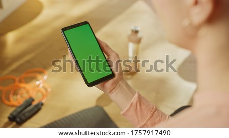 Fitness Concept: Woman Holding Chroma Key Green Screen Smartphone. In the Background Fitness Mat, Jumping Rope and Equipment. Close-up Over The Shoulde Shot.