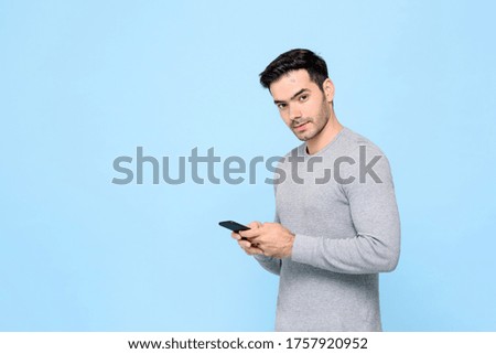 Portrait of handsome Caucasian man texting message on mobile phone and looking at camera in isolated studio blue  background