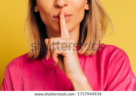 Cut view picture of young blonde woman holding finger upon lips in hush sign. Keep secret or word in mouth. Calm peaceful model in pink shirt. Isolated over yellow background