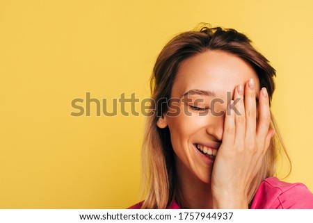 Picture of young blonde woman smiling and posing on camera. Girl keep half of face part with hand and smile. Mysterious model laughing and posing. Isolated over yellow background