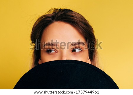 Close up picture of mysterious young woman posing on camera. Hide mouth behind black hat and look left. Beautiful stranger isolated over yellow background