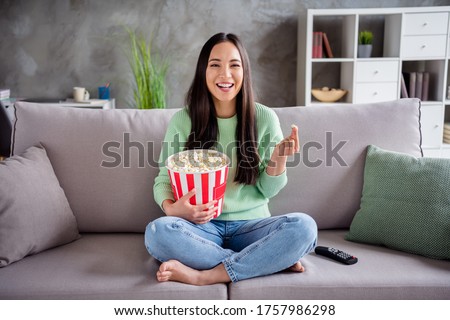 Full size photo of charming candid girl asian have free time sit divan legs bare foot crossed watch funny series eat pop corn box laughing in house indoors