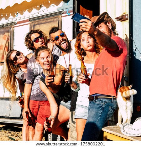 Group of alternative and diverse millennial people enjoy and have fun together taking selfie picture with phone and laughing like crazy in outdoor activity with old caravan in background