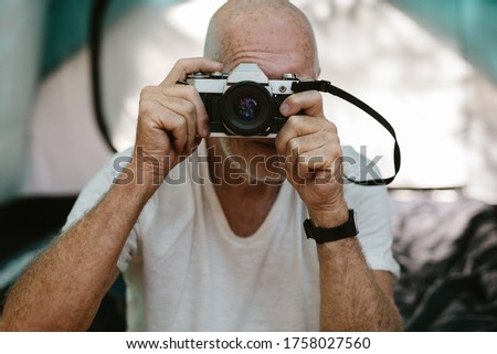 Closeup of a senior man sitting outside his camping tent and photographing with a digital camera. Retired man taking some snaps while camping in nature.