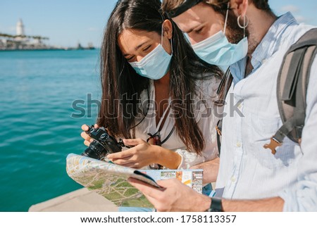 Young couple with a protective mask on their face looking at a map with a camera in their hand in front of the sea in a port