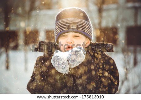 Little boy blows snow with mittens on bokeh background of snowflakes.