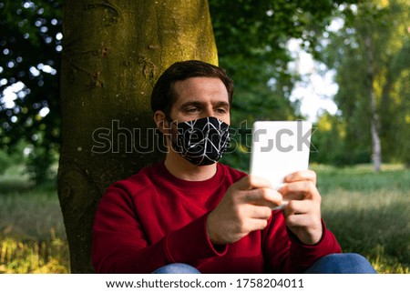 Photo of a young and attractive man sitting in the park reading an electronic book or ebook and wearing a reusable face mask


