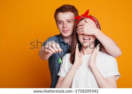 Red haired man closes eyes to the girl with his palm and holds keys to new car in front of her. Guy makes a surprise to his lover