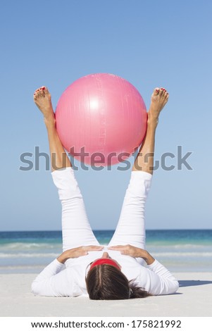 Portrait active and fit attractive looking mature woman at beach doing stretch exercises with gymnastic ball, legs up in the air, ocean and blue sky as blurred background.