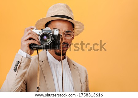 Snapshot of guy in stylish hat and jacket holding retro camera. Dark-skinned man in white shirt taking photo on isolated orange backdrop