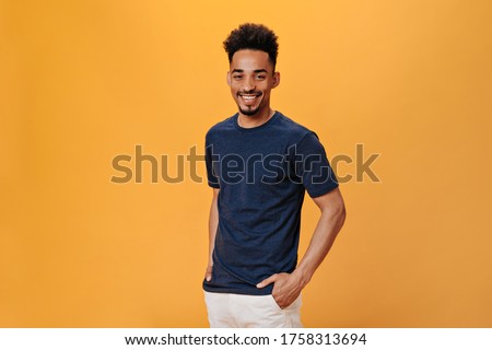 Man in black t-shirt smiles sweetly on orange background. Portrait of guy in blue tee posing on isolated backdrop
