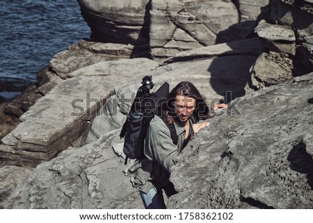 young long-haired guy with a backpack and a photo tripod climbs on a steep rock on the seashore. Climbing and outdoor concept.