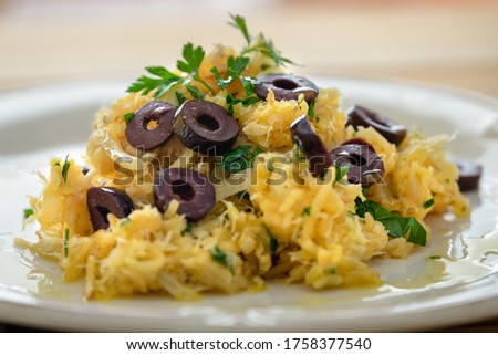 A typical Portuguese dish of shredded cod, with potato straw, mixed egg and dark olives. On a wooden table, with garlic, onion, parsley and others.