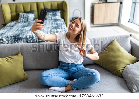 Portrait of attractive woman take selfie on smart phone gesturing v-sign with hand using cellphone enjoying video-call sitting indoor