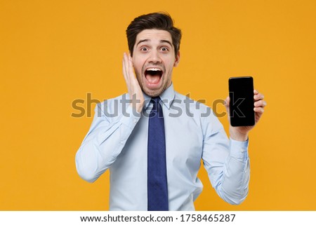 Shocked young business man in classic blue shirt tie isolated on yellow background studio. Achievement career wealth business concept. Hold mobile phone with blank empty screen, put hand on cheek