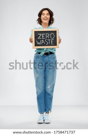 eco living, environment and sustainability concept - portrait of happy smiling young woman in turquoise shirt and jeans holding chalkboard with zero waste words grey background