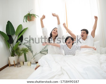 Happy family wake up in morning stretching hand rise up to the air while sitting on bed in bedroom with big window in background.Caucasian young man with asian woman and little boy exercise on bed. Royalty-Free Stock Photo #1758490187