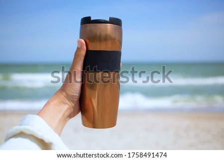 Woman hand hold bronze metal thermos cup 300 ml with coffee tea morning beverage during beautiful vacation at sea beach, enjoy drinking with view  Royalty-Free Stock Photo #1758491474
