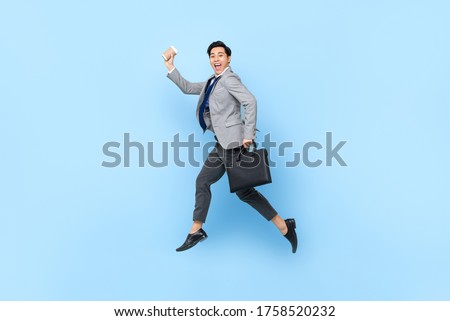 Happy cheerful young Asian businessman jumping while holding bag and coffee cup in isolated studio blue background
