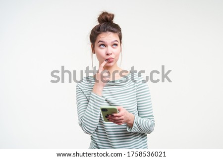 Thoughtful young pretty brown haired female with bun hairstyle keeping forefinger on her lip while looking pensively upwards, posing over white background