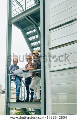 Three ambitious construction engineers standing on fire-escape of new building holding paper plan looking away, copy space Royalty-Free Stock Photo #1758542804