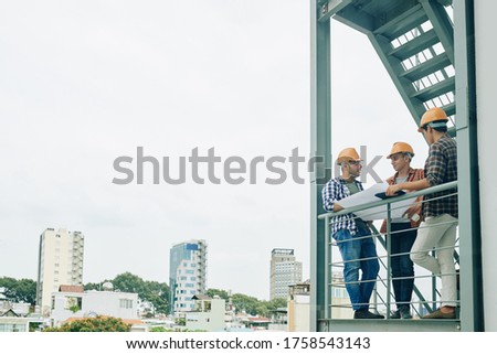 Group of three young construction engineers standing on fire-escape of new building holding paper plan and talking, copy space Royalty-Free Stock Photo #1758543143