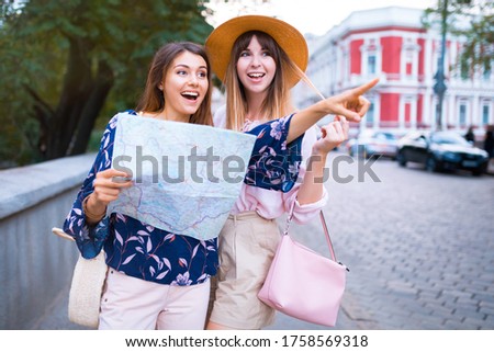 Happy travel together of two fashionable girls in sunny city centre. Young joyful women expressing positivity, using map, vacation with bags, camera, making photo, cheerful emotions, great mood