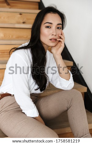 Smiling attractive young asian woman wearing white shirt sitting on a staircase at home