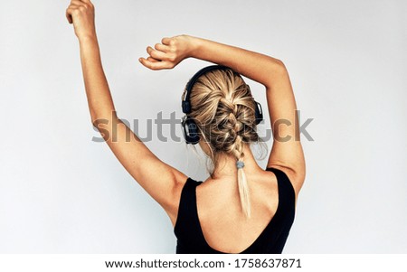 Beautiful back view image of attractive blonde young woman dancing during listening to music in headphones. Pretty female in black dress with braid hair listens to favourite songs in headphones