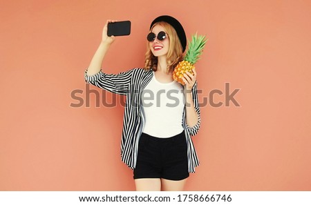 Summer portrait of happy smiling woman with pineapple taking selfie picture by smartphone on wall background