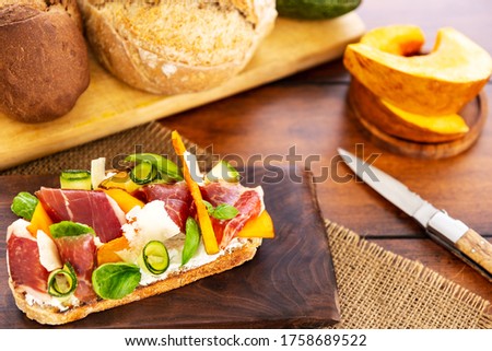 Open faced sandwich with iberico ham, parmesan and goat cheese, zucchini, pumpkin, basil and chives on sourdough bread. On a chopping board and wood, aside bread loaf, knife and pumpkin pieces.