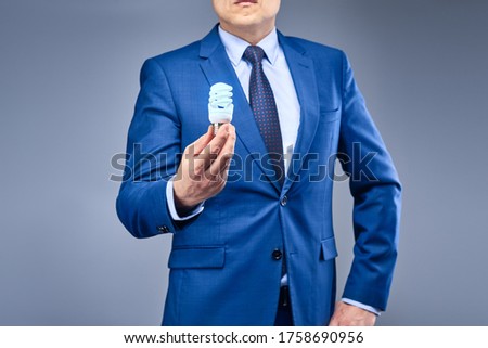A businessman in a blue suit on a gray-blue background holds a light bulb in front of him burning blue. The concept of finding an idea, making the right decision.