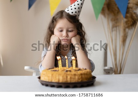 Cute baby on his birthday sits at the table propping his head with two fists. In front of her is a cake with candles. She has a birthday and no mood. The child is upset and sad.