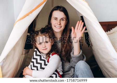 Happy young mom and cute little kid child daughter sitting in cozy tent talking video calling on chatting laughing enjoy online conversation looking at webcam, family vloggers recording video