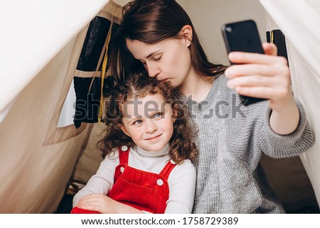 Happy young mom with cute kid daughter having fun holding smart phone sitting on cozy tent, mother kisses little girl recording vlog talking to webcam laugh embrace communicate online in internet