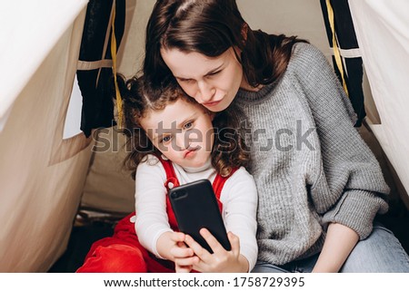 Cute little kid daughter and young mother funny face taking family picture look at smartphone, sitting in cozy tent, happy parent mum having fun with child girl make selfie together at home