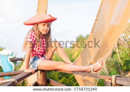 Serious funny little girl pirate in a plaid shirt and denim shorts holds on to the cords of the ship. Concept kids in adventure seekers