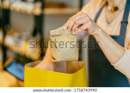 Hand of unrecognizable shopkeeper in package free grocery store collects paper bag of food.Food delivery service. Owner working behind counter at zero waste shop. Save planet, healthy life concept.