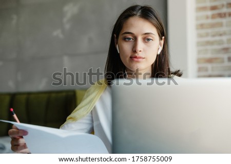 Attractive young brunette woman working on laptop computer while sitting at the table at home, taking notes