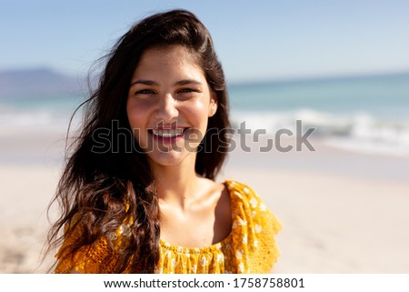Portrait of a Caucasian woman with long dark hair on holiday, enjoying free time on a sunny beach, looking to camera and smiling, with blue sky and sea in the background