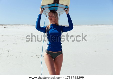 A Caucasian woman enjoying time at the beach on a sunny day, holding surfboard and walking, with blue sky in the background