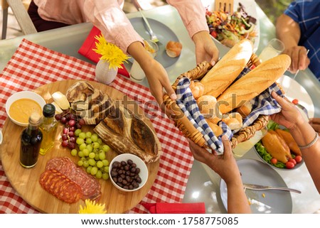 A mixed race woman passing a basket of bread across the table to her senior mother during a family meal outside on a patio in the sun, her father sitting at the end of the table in the background