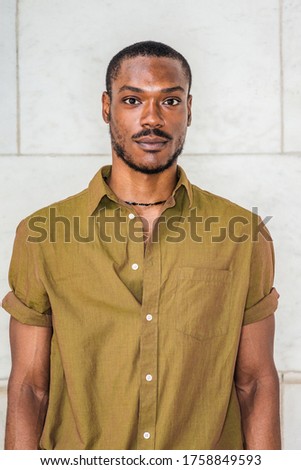 Close up, head short of Young African American Man with beard in New York, wearing green short sleeve shirt, necklace, collar unbuttoned, standing against white marble wall, seriously looking forward.