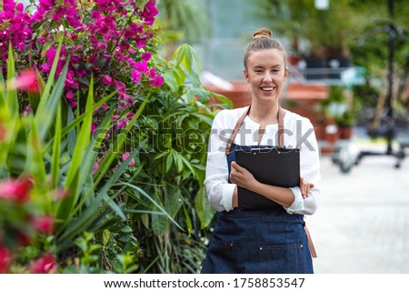 Young woman taking notes while working in a greenhouse. Woman working in a garden center and taking notes of her plants. Portrait of a young woman at work in greenhouse