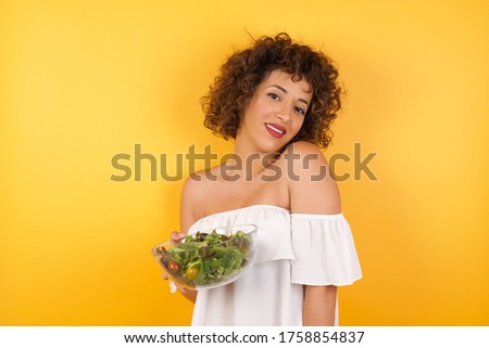 Shy girl holding a salad wants to start a new diet and lose weight, smiling looking to the camera. Caucasian charming girl sanding against yellow studio background feeling shy.