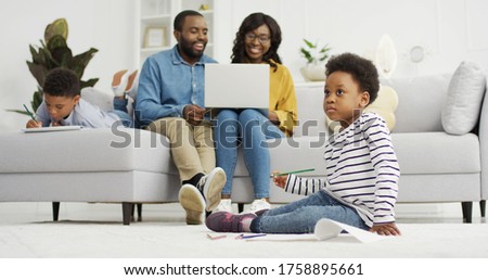 Happy african american family sitting on sofa together with device at home. Mother and father using laptop, small children brother and sister drawing at home, family time concept